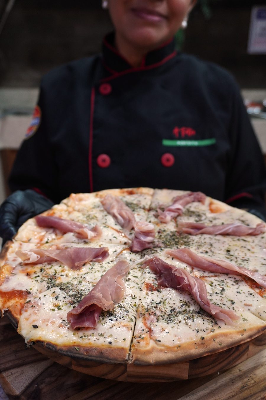 Integrante uniformizada da equipe da Fatia Perfeita segurando uma pizza de presunto de parma em tábua de madeira